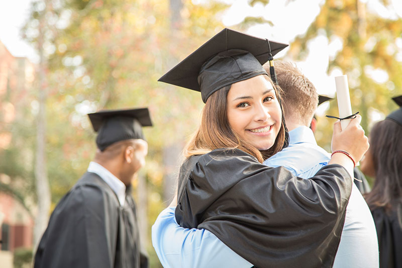 A school graduate dressed in cap and gown receiving a hug while holding her diploma