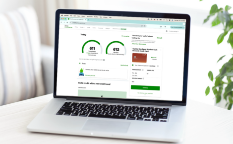 An image of a MacBook laptop on a white table or desk, a plant is seen peeking on the right side of the image. The computer screen shows a Credit Karma account's dashboard.