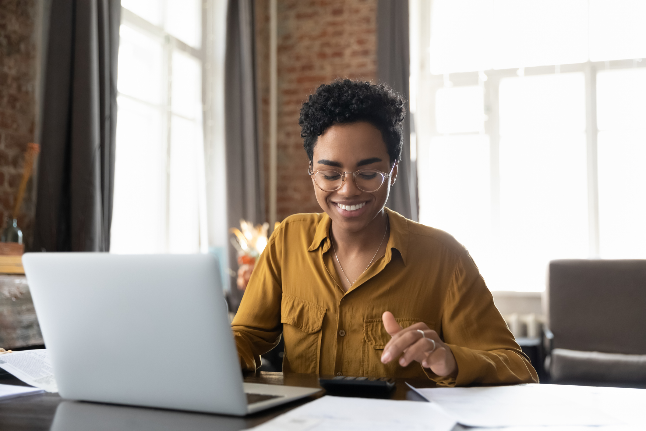 A smiling woman sits at her desk with her computer and a calculator to help her budget