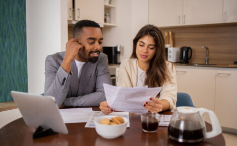 A young couple sitting at a kitchen table reviewing financial documents together, with a laptop, coffee, and a bowl of cookies in front of them.