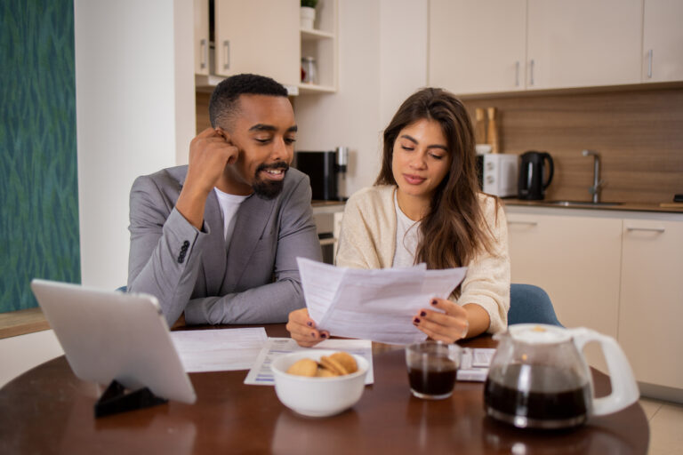 A young couple sitting at a kitchen table reviewing financial documents together, with a laptop, coffee, and a bowl of cookies in front of them.