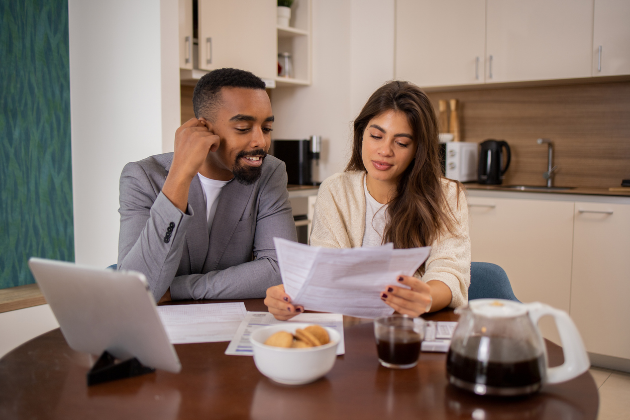 A young couple sitting at a kitchen table reviewing financial documents together, with a laptop, coffee, and a bowl of cookies in front of them.