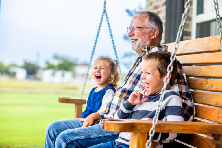 A grandfather sits on a porch swing in between his young grandson and young granddaughter, all laughing at a joke Grandpa told