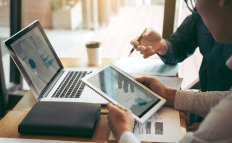 Two professionals reviewing financial data on a tablet and laptop during a meeting at a modern office desk.