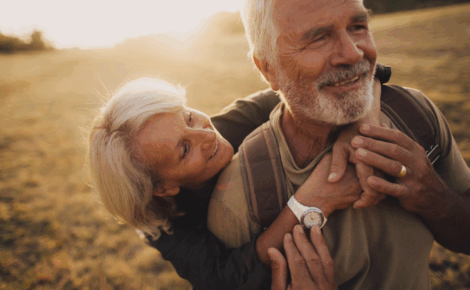 Smiling senior couple outdoors in a large field at sunset, with the woman embracing the man from behind as they walk together.
